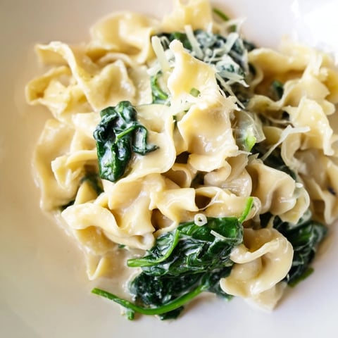 A close-up photo of creamy spinach garlic noodles twirling around a fork, with wilted green spinach leaves clinging to tender egg noodles in a rich, garlicky cream sauce.