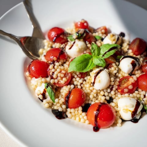 Freshly cooked pearled couscous salad with halved cherry tomatoes, creamy mozzarella balls, and torn basil leaves, all drizzled with sweet balsamic glaze on a rustic platter.
